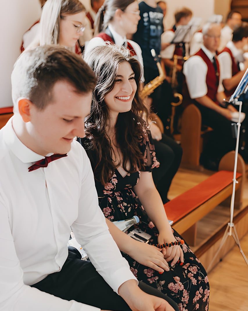 Dominik und Sophia sitzen nebeneinander in einer Kirchenbank und verfolgen lächelnd eine Trauung