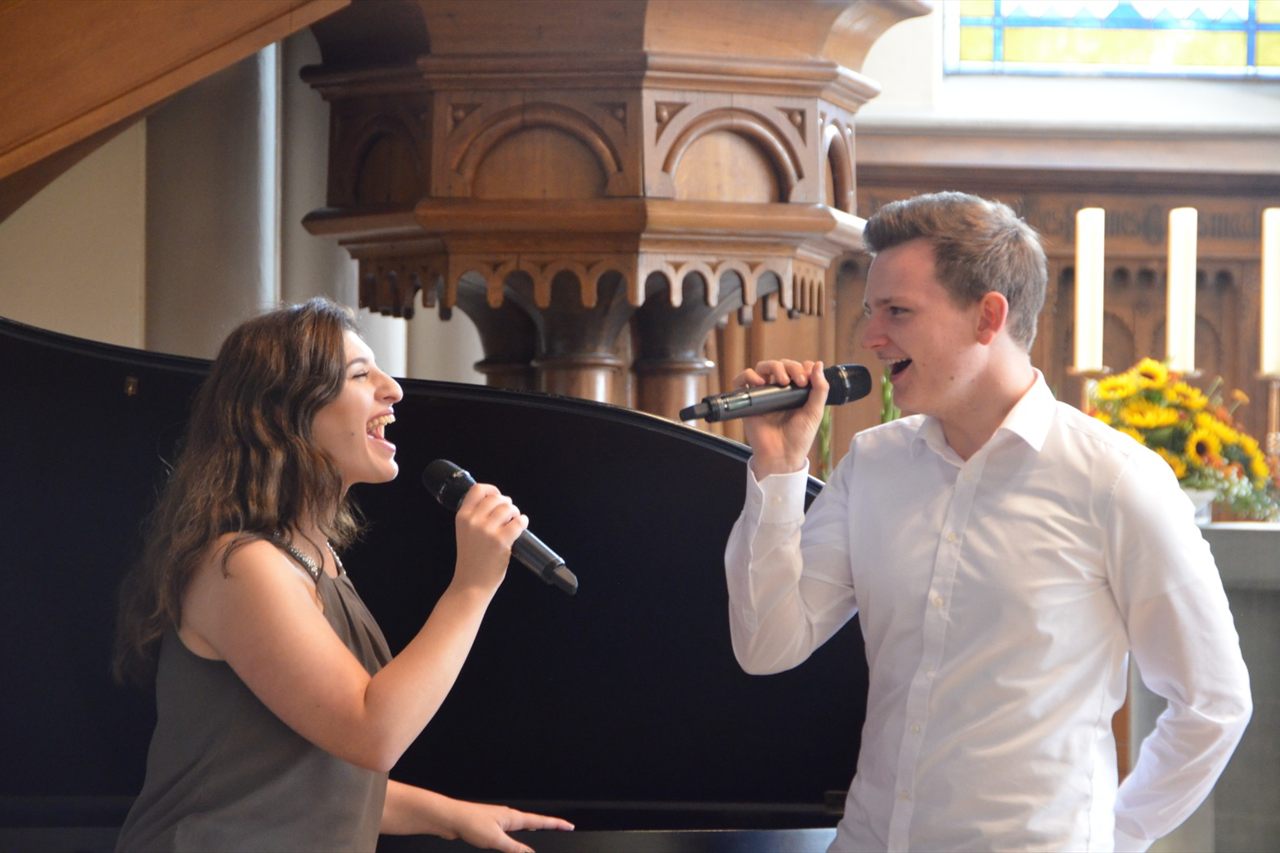 Dominik und Sophia singen gemeinsam mit Mikrofonen neben einem Flügel in einer Kirche